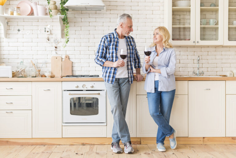 portrait-senior-couple-holding-wineglasses-hand-looking-each-other-standing-kitchen